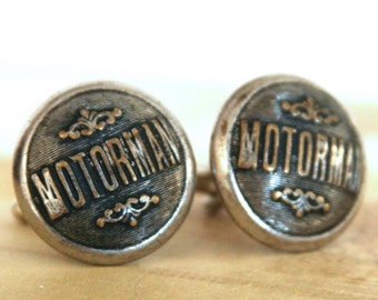 Puede incluir: Un par de gemelos vintage con un diseño circular. Los gemelos son metálicos con la palabra "MOTORMAN" en una fuente oscura. Hay adornos decorativos encima y debajo del texto. El fondo es de color marrón claro.