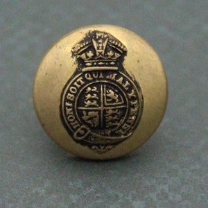 Puede incluir: Un botón dorado con un escudo real negro que presenta una corona y las palabras "Honi Soit Qui Mal Y Pense".