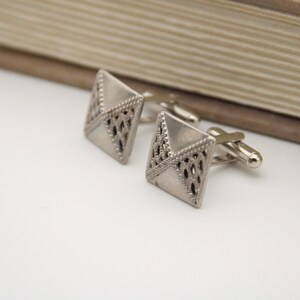 May include: A pair of silver cufflinks with a geometric design. The cufflinks are square shaped with a detailed pattern.