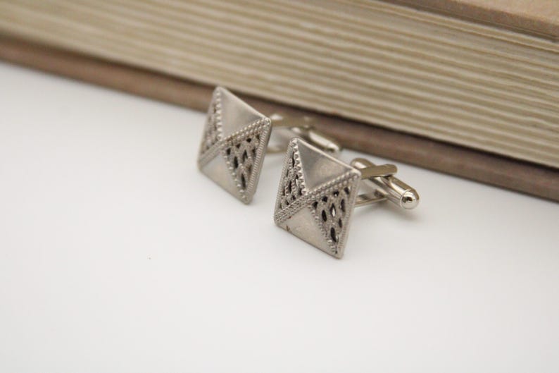 May include: A pair of silver cufflinks with a geometric design. The cufflinks are square shaped with a detailed pattern.