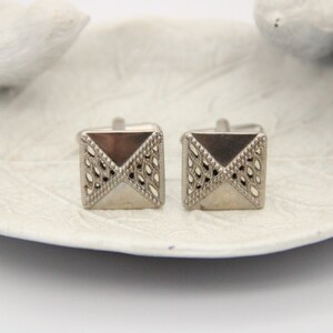 May include: A pair of silver cufflinks with a geometric design. The cufflinks are square shaped with a diamond pattern.