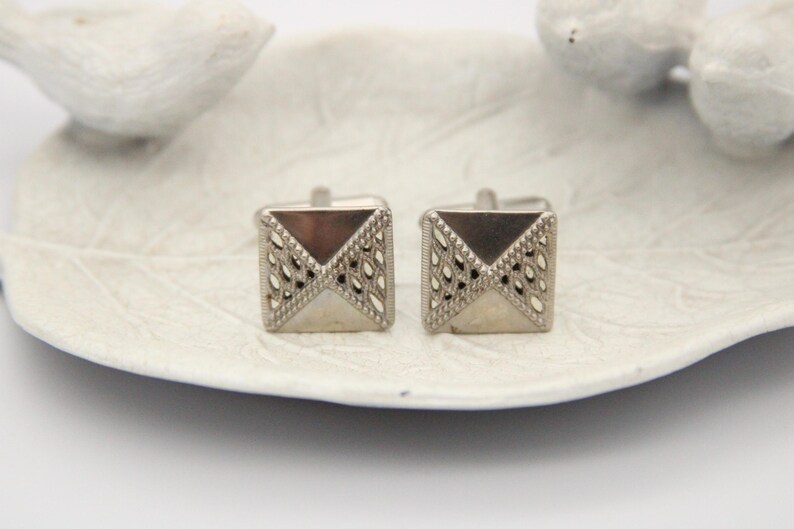 May include: A pair of silver cufflinks with a geometric design. The cufflinks are square shaped with a diamond pattern.