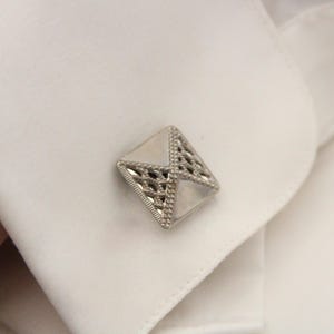May include: A silver cufflink with a geometric design. The cufflink is made of metal and has a shiny finish.
