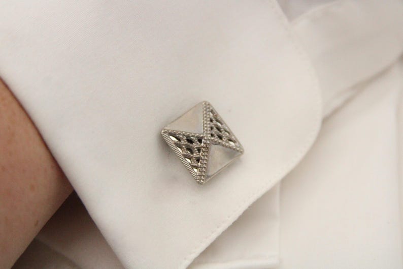 May include: A silver cufflink with a geometric design. The cufflink is made of metal and has a shiny finish.