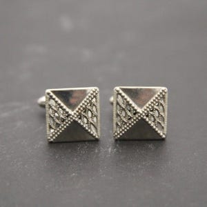 May include: A pair of silver cufflinks with a geometric design. The cufflinks are square shaped with a raised, triangular pattern.