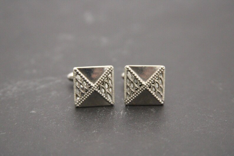 May include: A pair of silver cufflinks with a geometric design. The cufflinks are square shaped with a raised, triangular pattern.