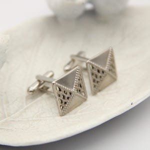 May include: A pair of silver cufflinks with a geometric design. The cufflinks are square shaped with a diamond pattern.