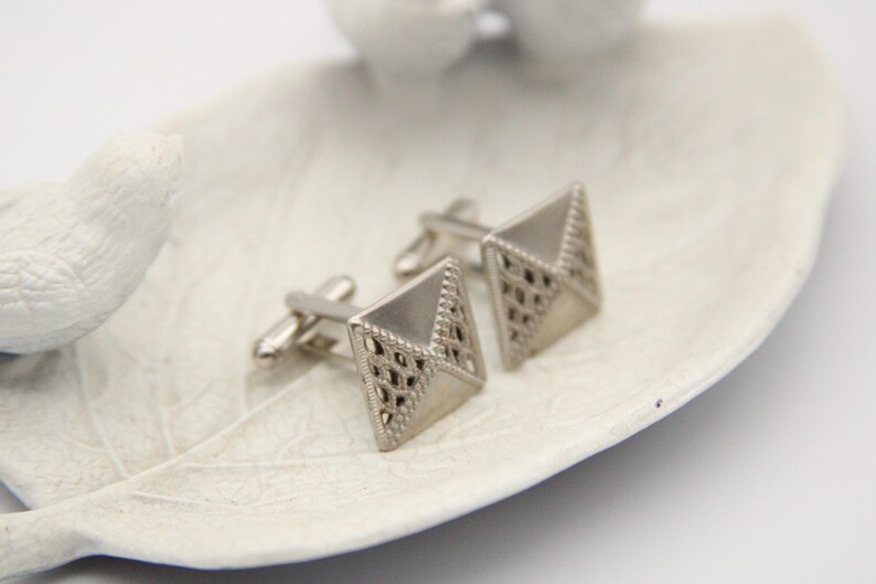 May include: A pair of silver cufflinks with a geometric design. The cufflinks are square shaped with a diamond pattern.