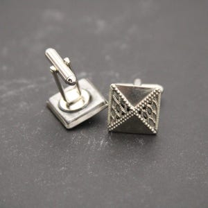 May include: A pair of silver cufflinks. One cufflink has a geometric design and the other has a simple, square shape.