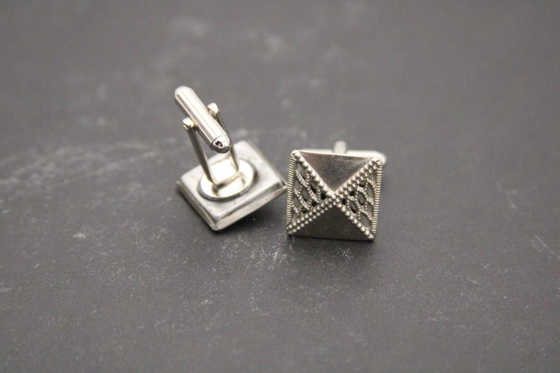 May include: A pair of silver cufflinks. One cufflink has a geometric design and the other has a simple, square shape.