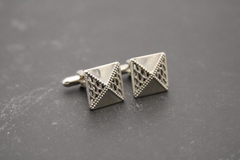 May include: A pair of silver cufflinks with a geometric design. The cufflinks are shaped like pyramids with a textured surface.