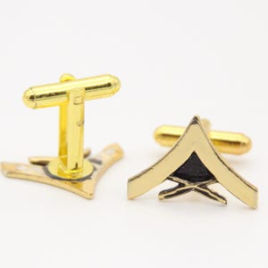 May include: A pair of gold-toned metal cufflinks. One cufflink is a T-shape, and the other is a gold chevron with a black center. The cufflinks are on a white background.