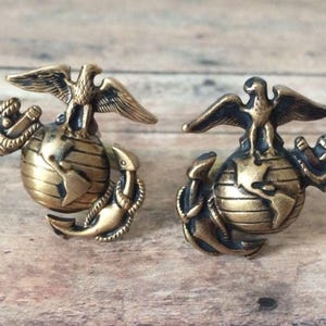 May include: Two gold-toned metal cufflinks featuring the United States Marine Corps emblem. Each cufflink depicts an eagle perched atop a globe with an anchor below.