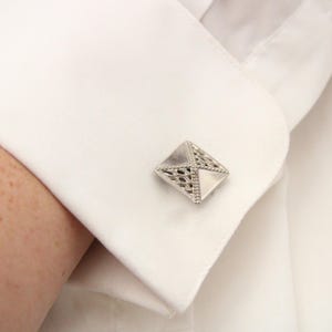 May include: A silver cufflink with a geometric design. The cufflink is a square shape with a raised, intricate pattern.