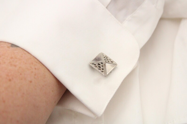May include: A silver cufflink with a geometric design. The cufflink is a square shape with a raised, intricate pattern.