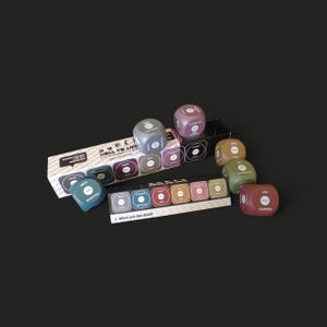 May include: A collection of multi-coloured dice, each bearing a different word such as "Flowers" and "Roastful." The dice are presented with a black box displaying the text "Roll to Life" and a card asking "What are the dice?" The dice are in shades of blue, green, brown, and pink.