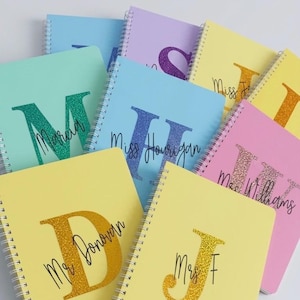 May include: A collection of spiral-bound notebooks in pastel colours including mint, light blue, yellow, lavender, and pink. Each notebook features a large glittery initial letter and a personalised name printed in black cursive script on the cover.