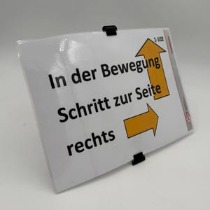 May include: A white sign with black text and yellow arrows, held by black clips. The text reads "In der Bewegung Schritt zur Seite rechts". The sign has a clear plastic cover and is angled towards the viewer.