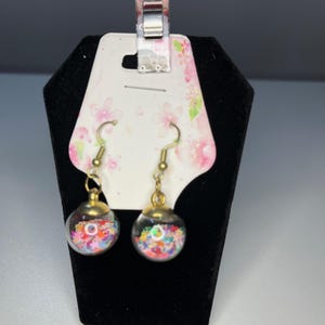May include: A pair of dangle earrings featuring clear, spherical charms filled with colorful confetti. The earrings have gold-colored metal accents and hooks. They are displayed on a black velvet earring stand with a floral-patterned tag.