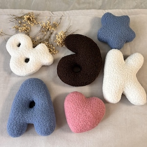 May include: A collection of plush, textured sherpa-style cushions in various shapes including the letters A, K, and an ampersand, a dark brown number 6, a blue star, and a pink heart, accompanied by a small cluster of dried baby's breath flowers.