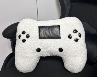 May include: A white, fluffy cushion in the shape of a games controller. It features black buttons and a black rectangular panel. The cushion is placed on a black chair.