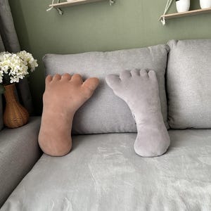 May include: Two foot-shaped cushions. One is light tan, the other light grey. They rest on a grey sofa. The cushions are a quirky home decor item. The cushions are a fun and unique home decor item.