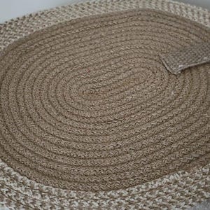 Op de afbeelding: Ovaal gevlochten jute tapijt in beige en bruin. Het midden is effen bruin, omgeven door een rand van afwisselend beige en bruin gevlochten strengen. Een klein, rechthoekig stuk van het tapijt is omgeslagen.
