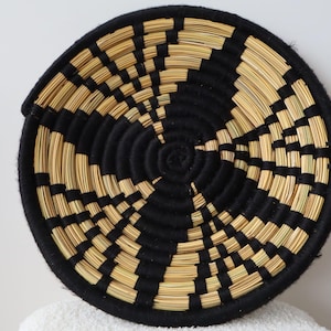 Op de afbeelding: Een ronde, gewikkelde wandmand met een geometrisch patroon van afwisselend zwart garen en natuurlijk bruin stro. Het ontwerp straalt vanuit het midden in een spiraalvorm, wat zorgt voor een contrastrijke, getextureerde uitstraling met een dikke rand.