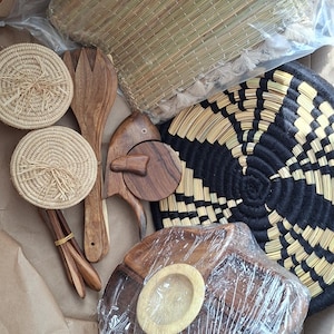 Op de afbeelding: Een verzameling huishoudelijke artikelen, waaronder een geweven strodmat in plastic, twee ronde geweven onderzetters, een houten slavork, een houten serveerstuk in de vorm van een vis, een zwart-beige geweven mand en een houten serveerblad in plastic.