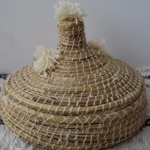 Op de afbeelding: Een ronde mand met deksel, gemaakt van lichtbruin gevlochten zeegras. Het deksel heeft een hoog, puntig handvat in het midden en drie decoratieve, gebroken witte garenkwastjes. De mand heeft een brede, platte basis en een spiraalstructuur.