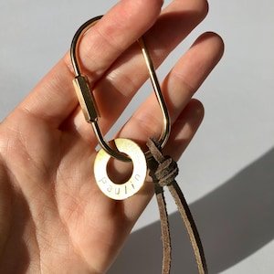 Könnte beinhalten: Ein goldfarbener Karabiner-Schlüsselanhänger mit einem kreisförmigen Anhänger mit der Aufschrift "paulin" in einer Serifenschrift. Der Schlüsselanhänger hat einen braunen Lederriemen. Der Karabiner wird in einer Hand gehalten.