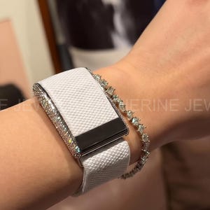 May include: A white textured wristband with a silver and diamond-studded frame and a black rectangular clasp. A diamond bracelet and a diamond ring are also visible. The image is of a jewelry product.