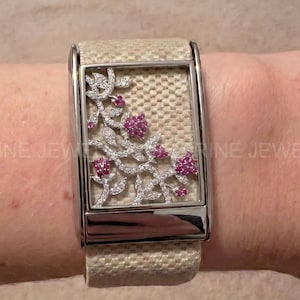 May include: A rectangular watch face featuring a textured beige fabric background with a silver-toned metal frame. The watch face is decorated with a silver vine pattern accented by small pink stones. The watch band is made of a matching beige fabric.