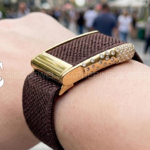 May include: A dark brown elastic watch band featuring a gold-toned metal connector piece. The connector is embellished with a series of small, clear, round crystals arranged in a scattered pattern across the surface of the gold metal hardware.
