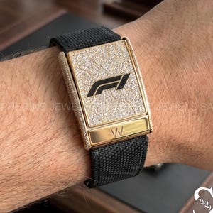 Könnte beinhalten: Eine rechteckige goldene Uhr mit schwarzem Stoffarmband. Das Zifferblatt hat eine mit Diamanten besetzte Oberfläche und ein schwarzes Formel-1-Logo in der Mitte. Ein glatter goldener Balken mit den Buchstaben W befindet sich unter dem Diamantbereich.
