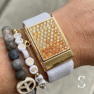 May include: A gold-toned rectangular watch with a white band, adorned with a gradient of orange and yellow gemstones. The watch is paired with beaded bracelets featuring gray and white beads, and the word "STRONG" spelled out in white letter beads.