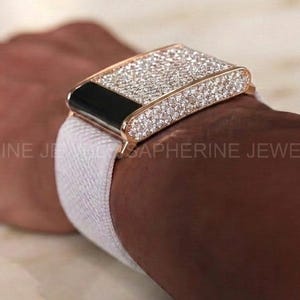 May include: A rectangular smartwatch case cover featuring a dense pavé setting of clear, sparkling stones in a metallic frame, paired with a plain white elastic fabric watch band. The device has a black screen section and a metallic gold-tone finish.