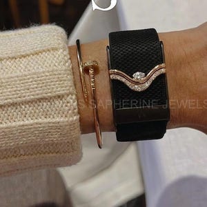 May include: A black textured watch band featuring a rose gold-toned V-shaped decorative piece adorned with small clear stones. Beside it is a thin rose gold-toned nail-shaped bangle bracelet. The watch band has a subtle, repeating pattern.