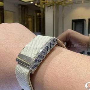 May include: A white fabric bracelet with a rectangular silver clasp. The clasp is adorned with small blue and clear gemstones. The bracelet is on a wrist.