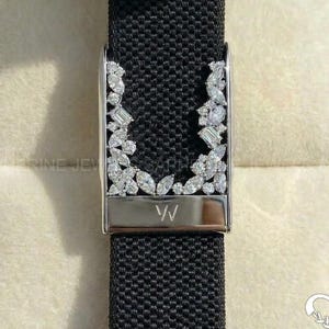 May include: A black textured watch strap featuring a silver-toned metal buckle adorned with a cluster of clear, faceted stones arranged in a U-shape. The metal component is engraved with the letters VV at the bottom center of the piece.