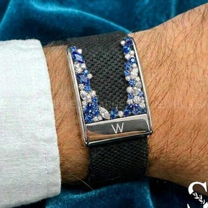 May include: A black fabric watch strap featuring a U-shaped metal accent adorned with blue and clear marquise and round-cut stones. A polished silver-tone metal bar at the base is engraved with the letter W, centered on the textured black band.
