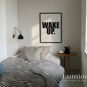 May include: A framed wall art print featuring the black text WAKE UP. on a white background, displayed above a bed with black and white striped bedding, a small wooden side table with a black pitcher and white cup, and a gold wall lamp.