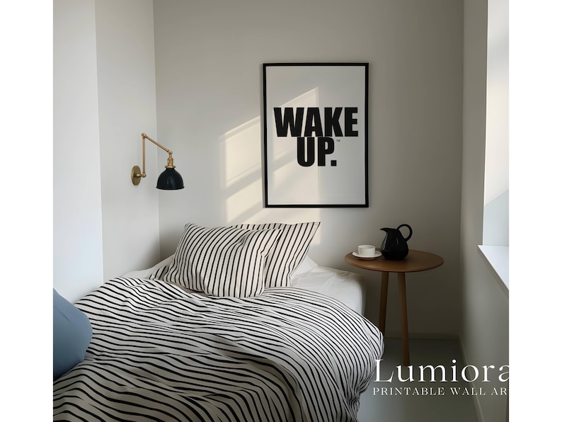 May include: A framed wall art print featuring the black text WAKE UP. on a white background, displayed above a bed with black and white striped bedding, a small wooden side table with a black pitcher and white cup, and a gold wall lamp.