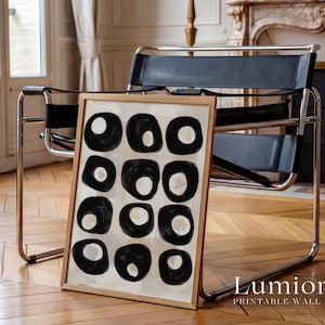 May include: A framed rectangular wall art print featuring a grid of twelve black hand-painted circular shapes on a light beige background. The artwork is encased in a light-colored wooden frame. The text "Lumiora PRINTABLE WALL ART" is visible.