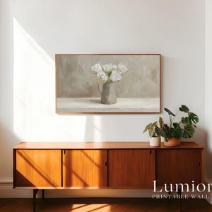 May include: A framed print of a still life painting featuring white flowers in a gray vase. The artwork hangs above a wooden cabinet with sliding doors. The cabinet is topped with potted plants. The text "Lumiora Printable Wall Art" is at the bottom.