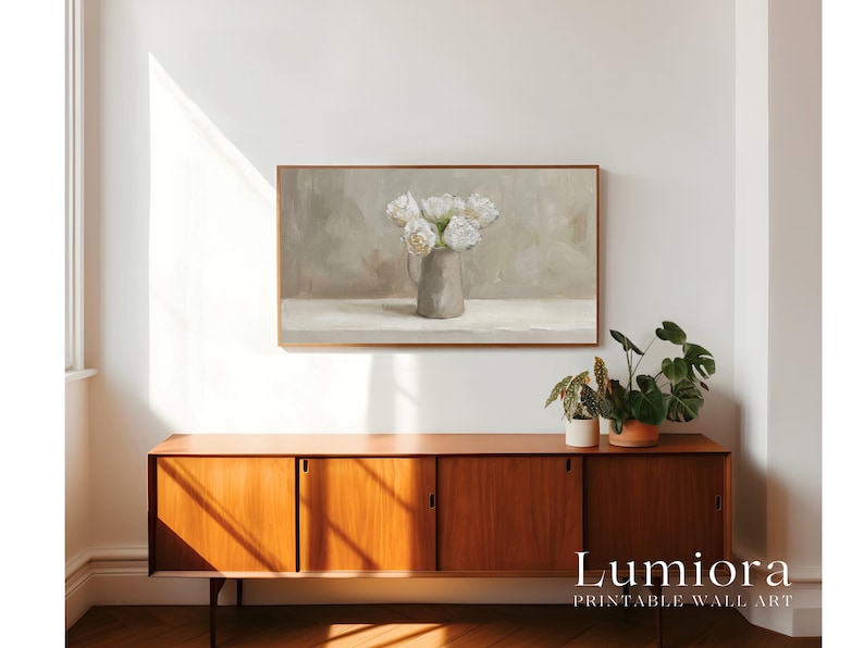 May include: A framed print of a still life painting featuring white flowers in a gray vase. The artwork hangs above a wooden cabinet with sliding doors. The cabinet is topped with potted plants. The text "Lumiora Printable Wall Art" is at the bottom.