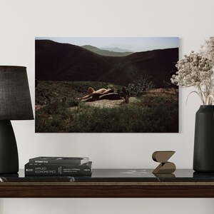 Puede incluir: Fotografía de una persona tumbada en un paisaje desértico, enmarcada por una lámpara negra, un jarrón con flores secas y una mesa de madera oscura. La escena evoca una sensación de tranquilidad y belleza natural.