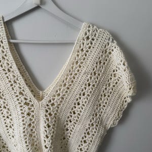 May include: Cream-colored, open-weave crochet top with a deep V-neck and short sleeves. The top is hanging on a white wooden hanger against a plain white wall. The crochet pattern features a delicate, airy design.