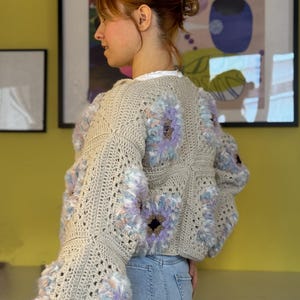 Op de afbeelding: Een beige gehaakte trui met grote, pluizige bloemaccenten in tinten paars, blauw en perzik. De trui heeft een los, open geweven ontwerp en klokmouwen. De persoon draagt een lichtblauwe spijkerbroek.