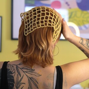 Pode incluir: Uma touca de croch&ecirc; bege com um design em forma de rede. A touca &eacute; usada na cabe&ccedil;a. A pessoa na imagem est&aacute; usando uma regata preta e tem uma tatuagem no bra&ccedil;o e nas costas.